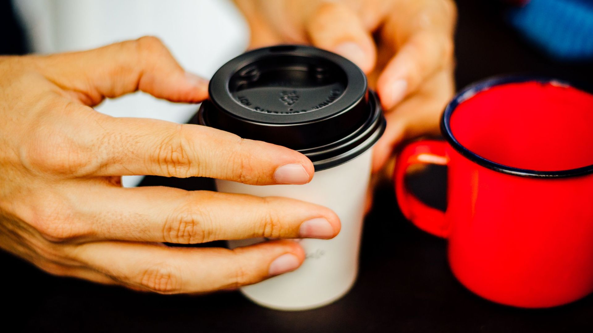 A close-up image of hands holding a disposable coffee cup with a plastic lid next to a red mug.