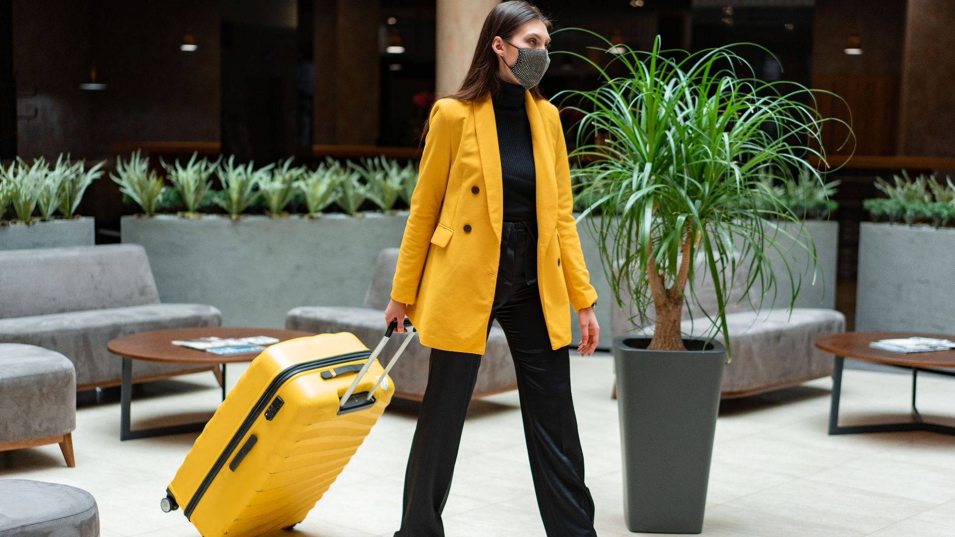 Fashionable woman in yellow blazer and face mask walking with suitcase indoors.