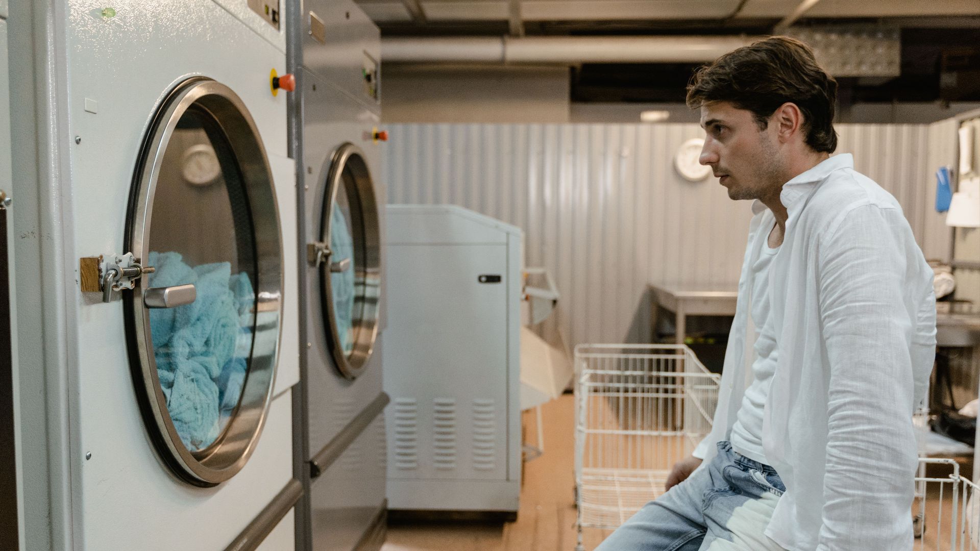 A man in a white shirt waits by machines in an industrial laundry facility.