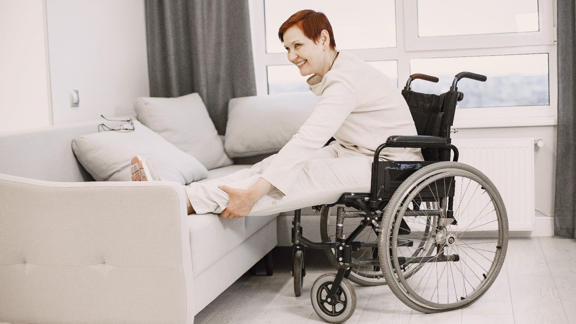 Smiling woman in wheelchair stretches legs on sofa in bright modern living room.