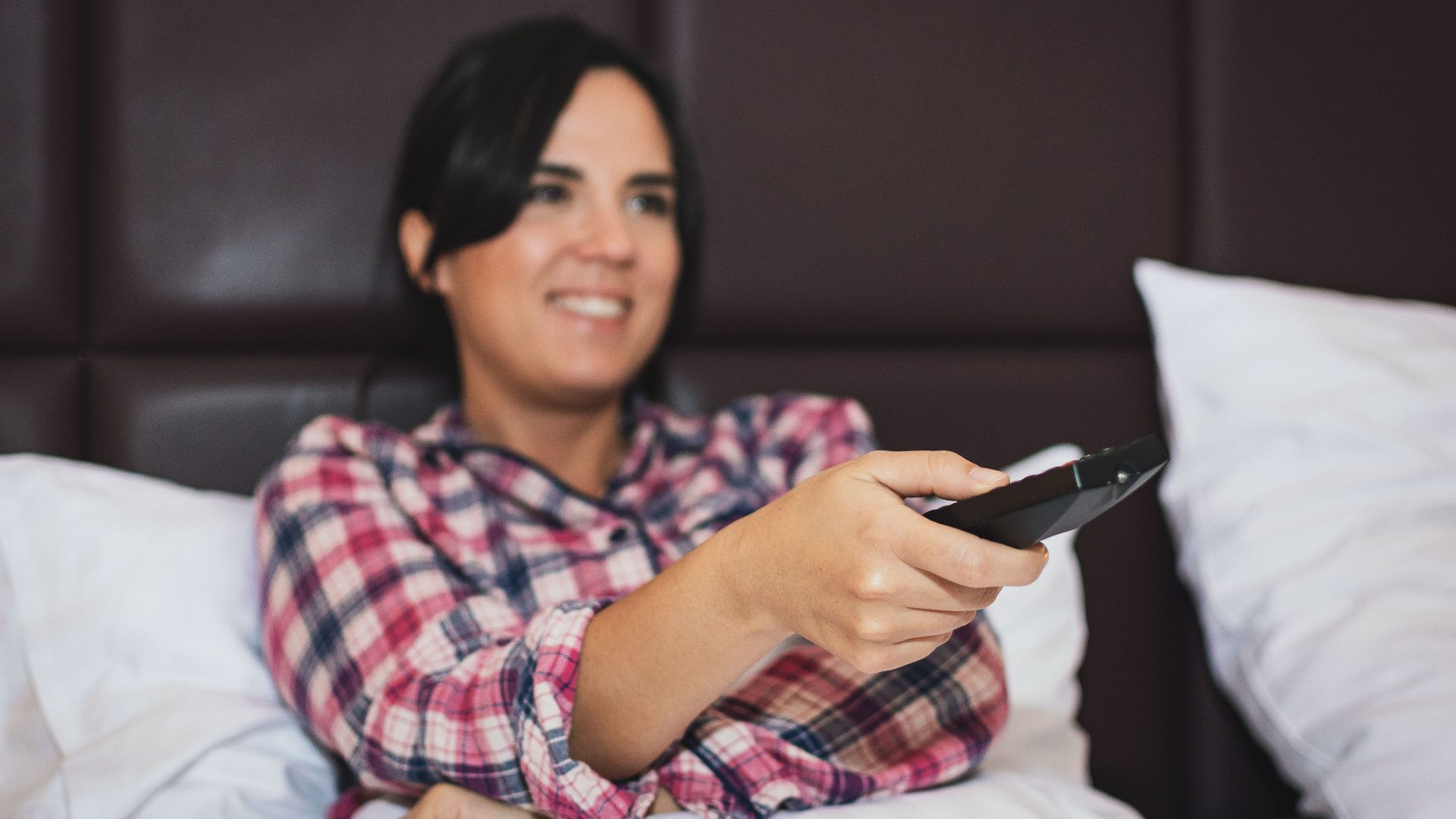 A woman in a plaid shirt smiling while holding a remote control in bed, enjoying leisure time.