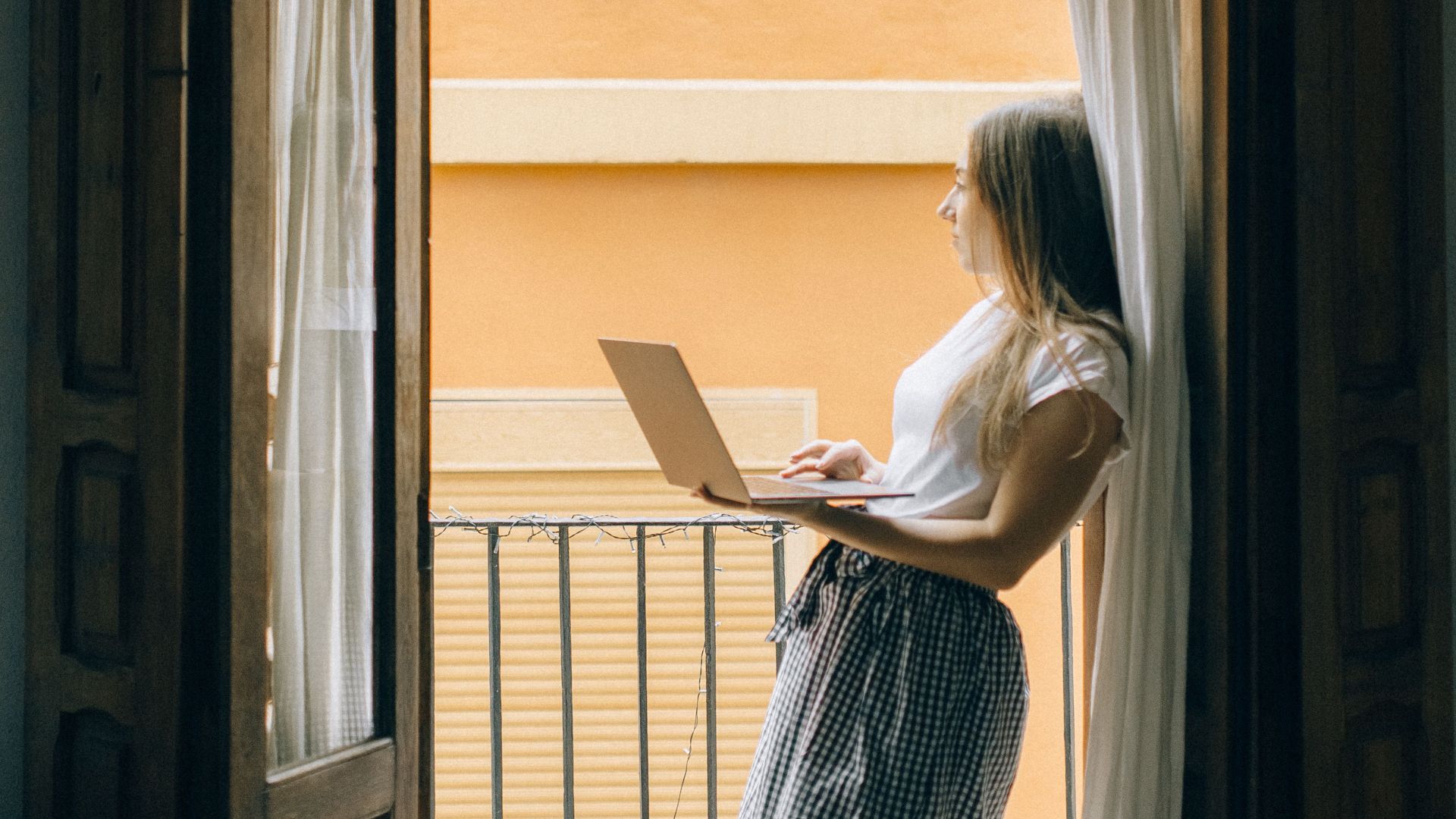 Woman standing barefoot on a balcony using a laptop, leaning against a wooden door frame.