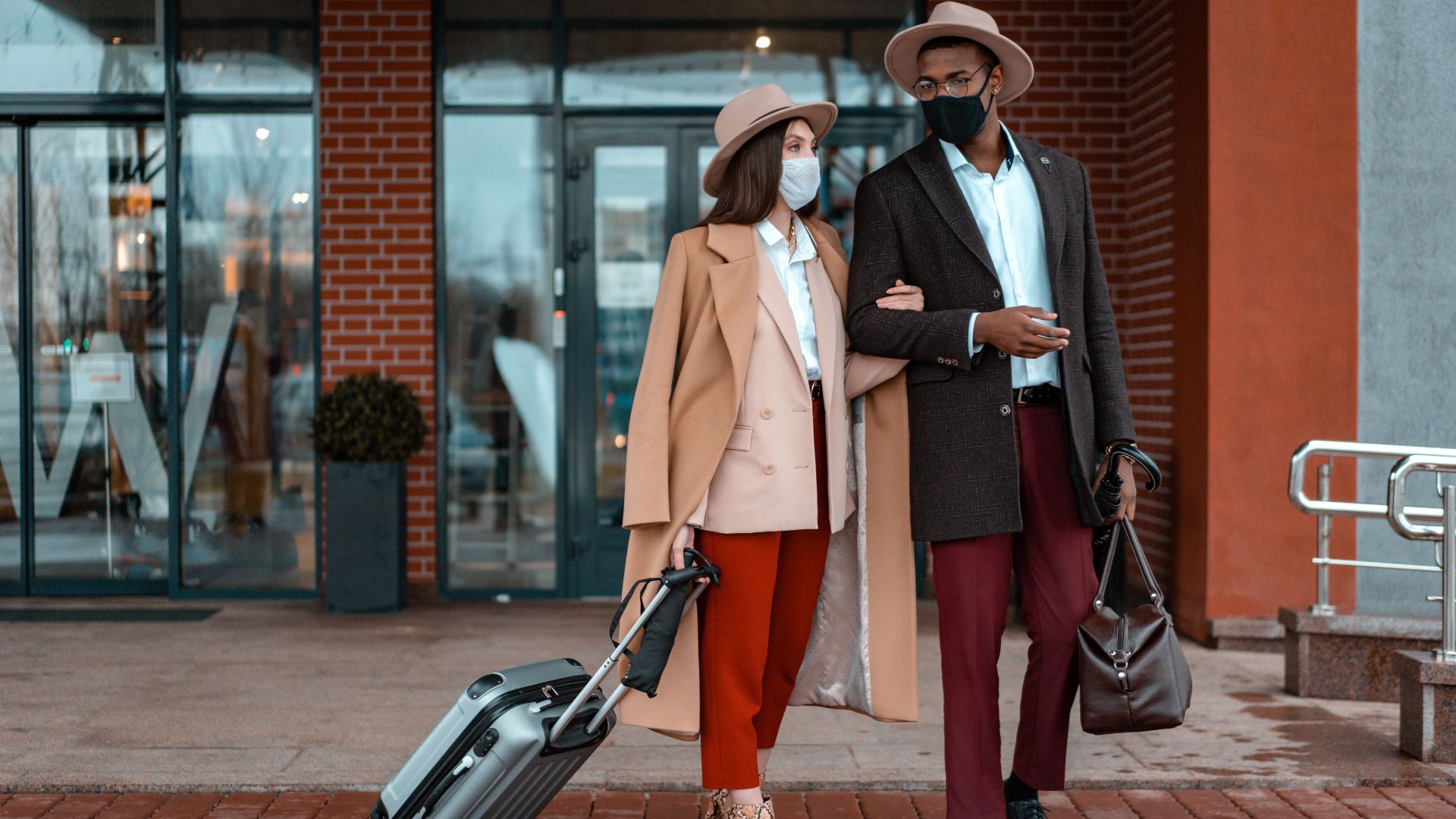 Fashionable couple with face masks at hotel entrance, showcasing travel during pandemic.