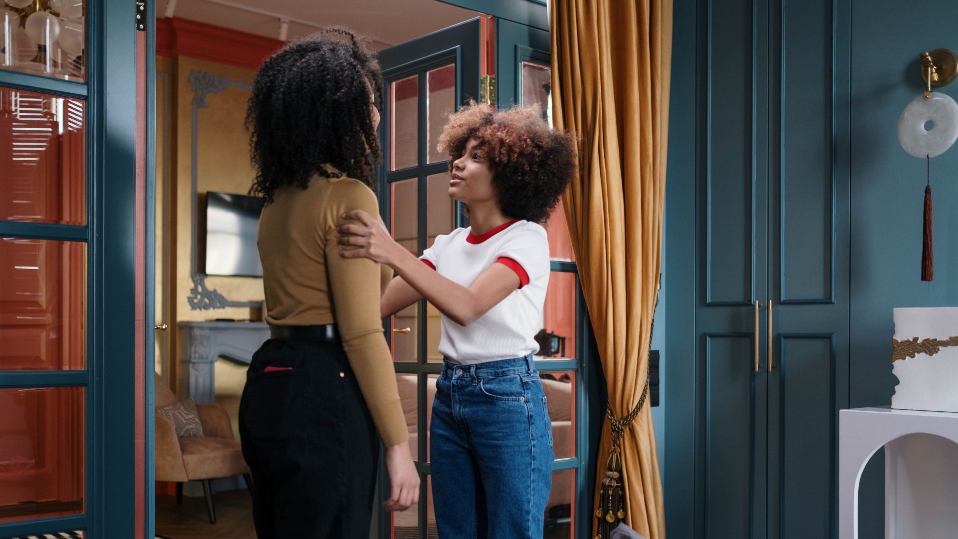 Two young women with curly hair share a moment of connection in a chic bedroom setting.