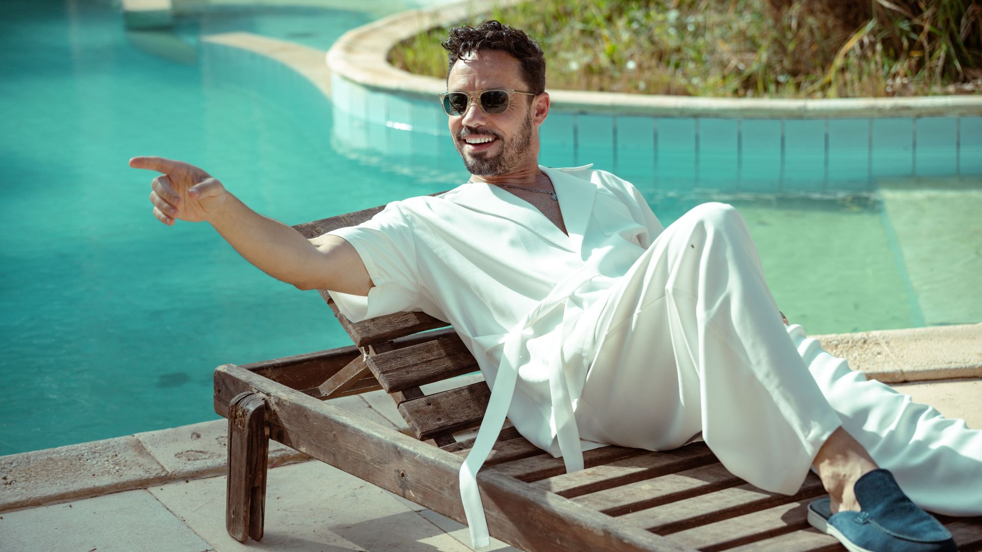 Man in white attire lounging on a sunbed by a pool, pointing and smiling.