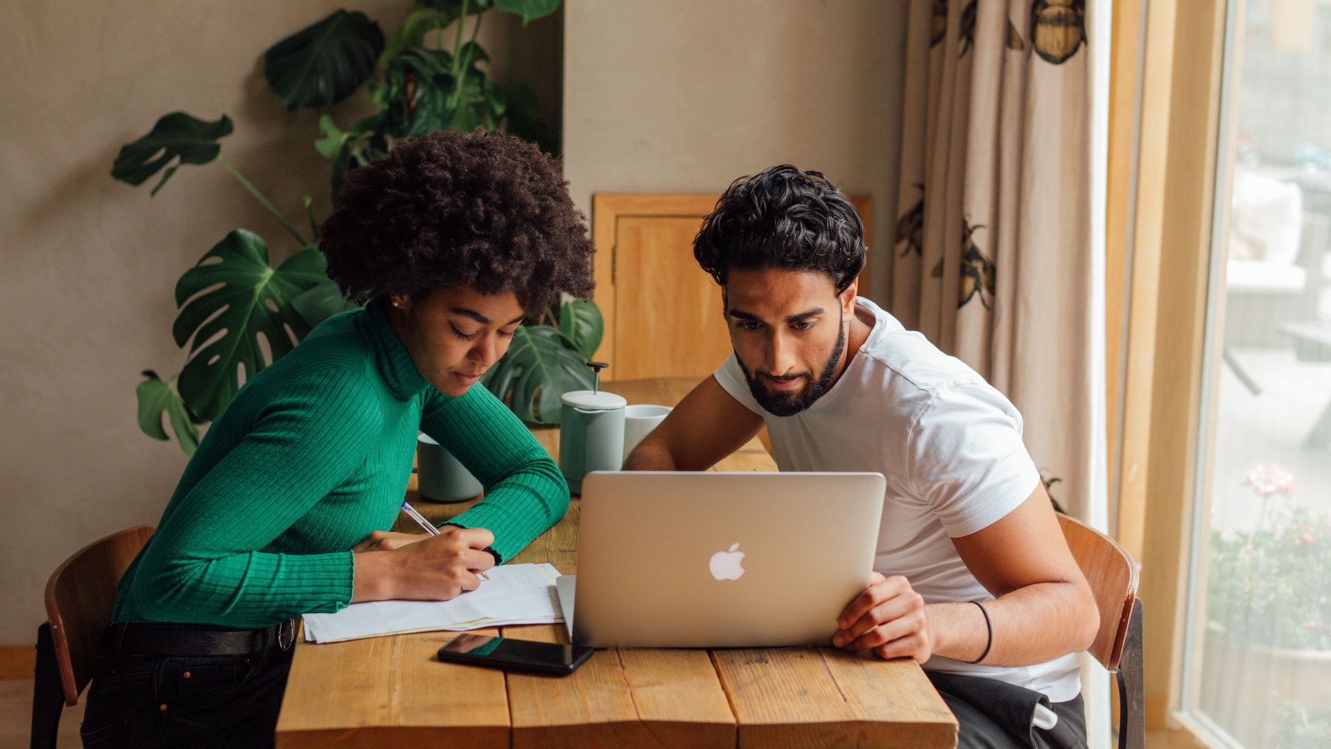 Two coworkers brainstorming and working together at a wooden table with a laptop indoors.