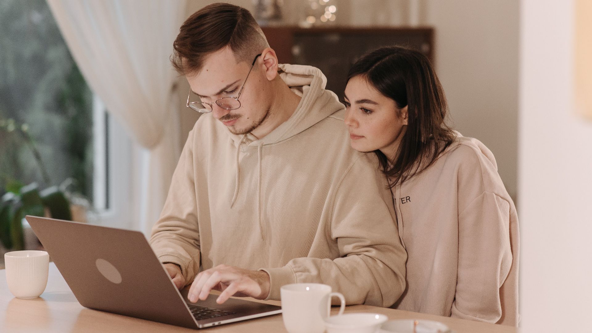 Young couple in hoodies using a laptop at home with coffee cups, embracing remote work lifestyle.