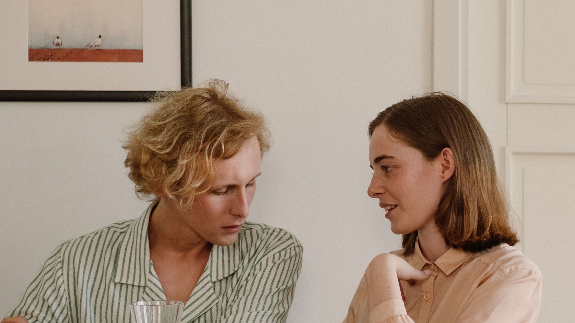 A couple engages in conversation by a piano, enjoying wine and books indoors.