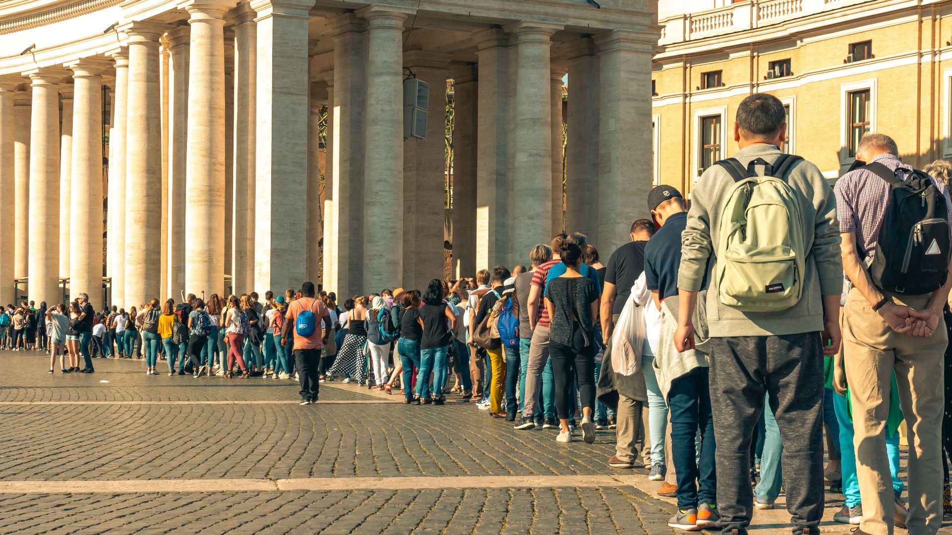 A large crowd of tourists lines up in sunny St. Peter's Square, showcasing architectural columns.