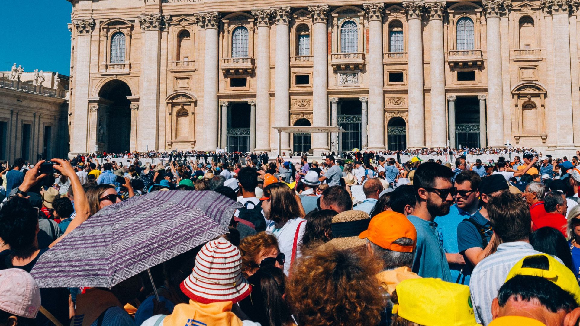 A large crowd gathers outside St. Peter's Basilica under a clear blue sky.