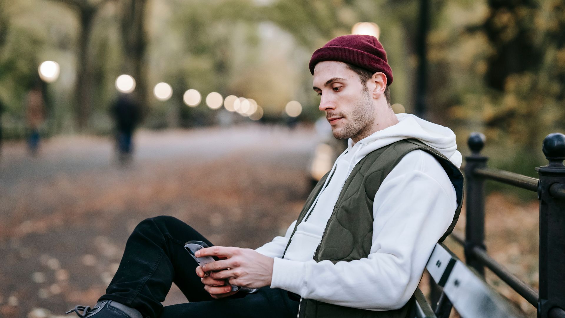 Side view of young unshaven male in casual wear and hat sitting on bench in autumn park and text messaging
