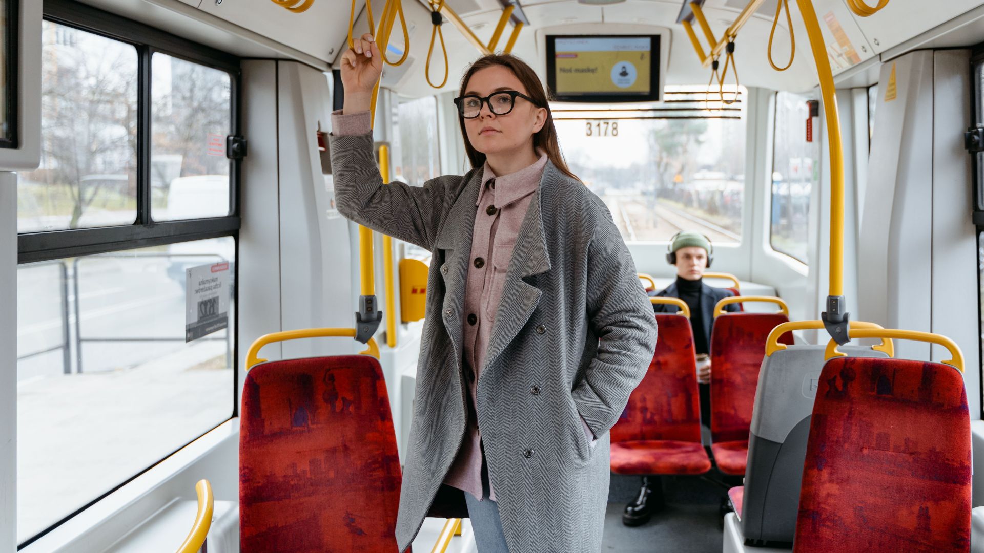A woman in a coat and eyewear standing inside a public tram, holding onto the handrail.