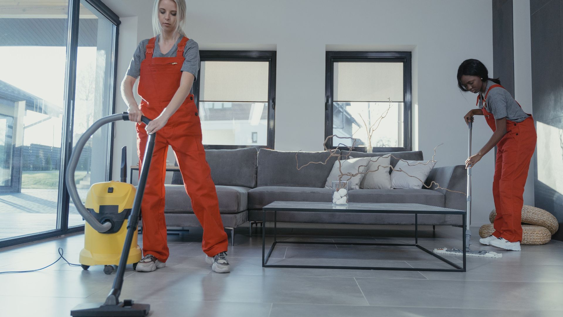 Two women in red overalls vacuuming and sweeping a modern living room.