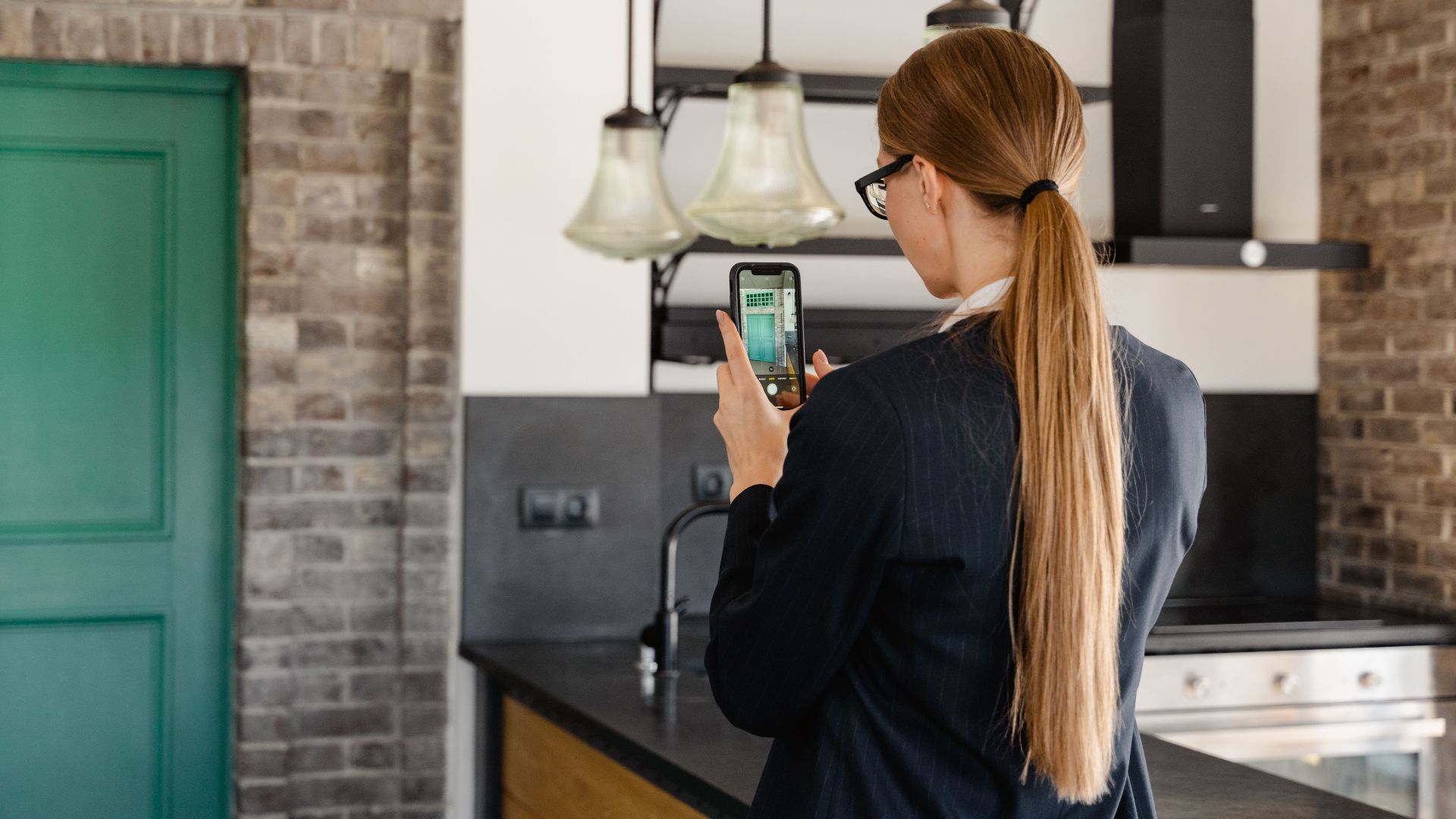 Rear view of a woman with long hair taking a photo with a smartphone in a stylish kitchen.
