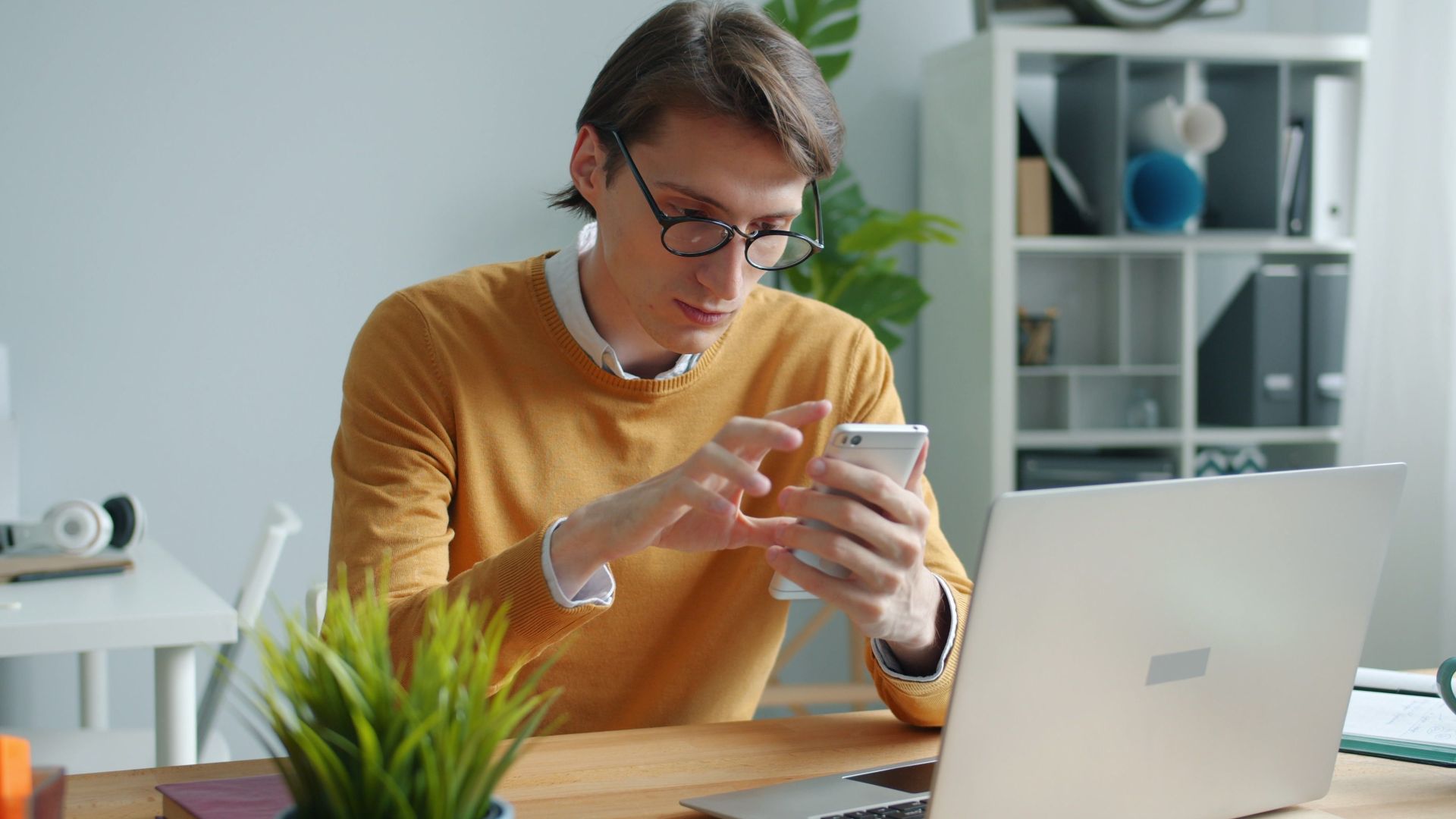 A young adult working on a smartphone and laptop in a modern office.