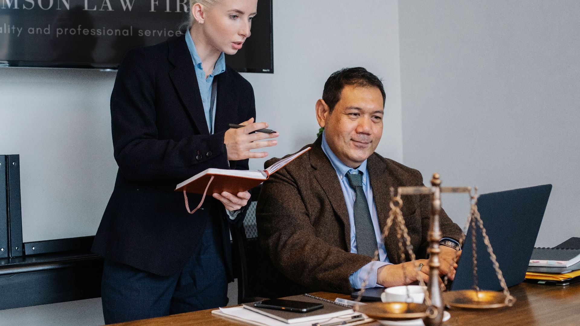 Concentrated young female assistant taking notes in planner while standing at table near middle aged ethnic male colleague typing on laptop during work in law firm