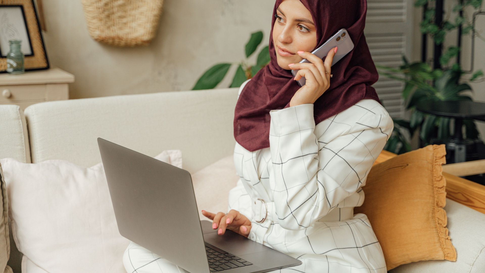 A Muslim woman in a hijab working remotely, balancing a phone call and laptop in a home office setting.