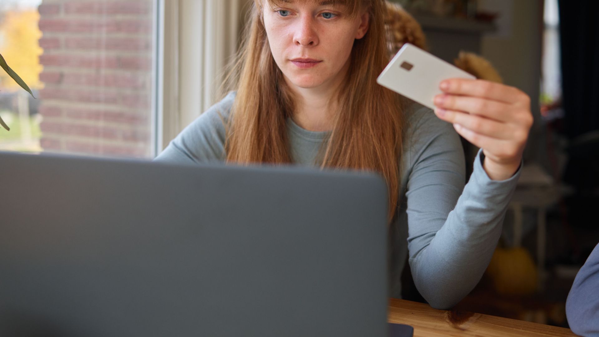 A young woman holds a credit card while using her laptop indoors.