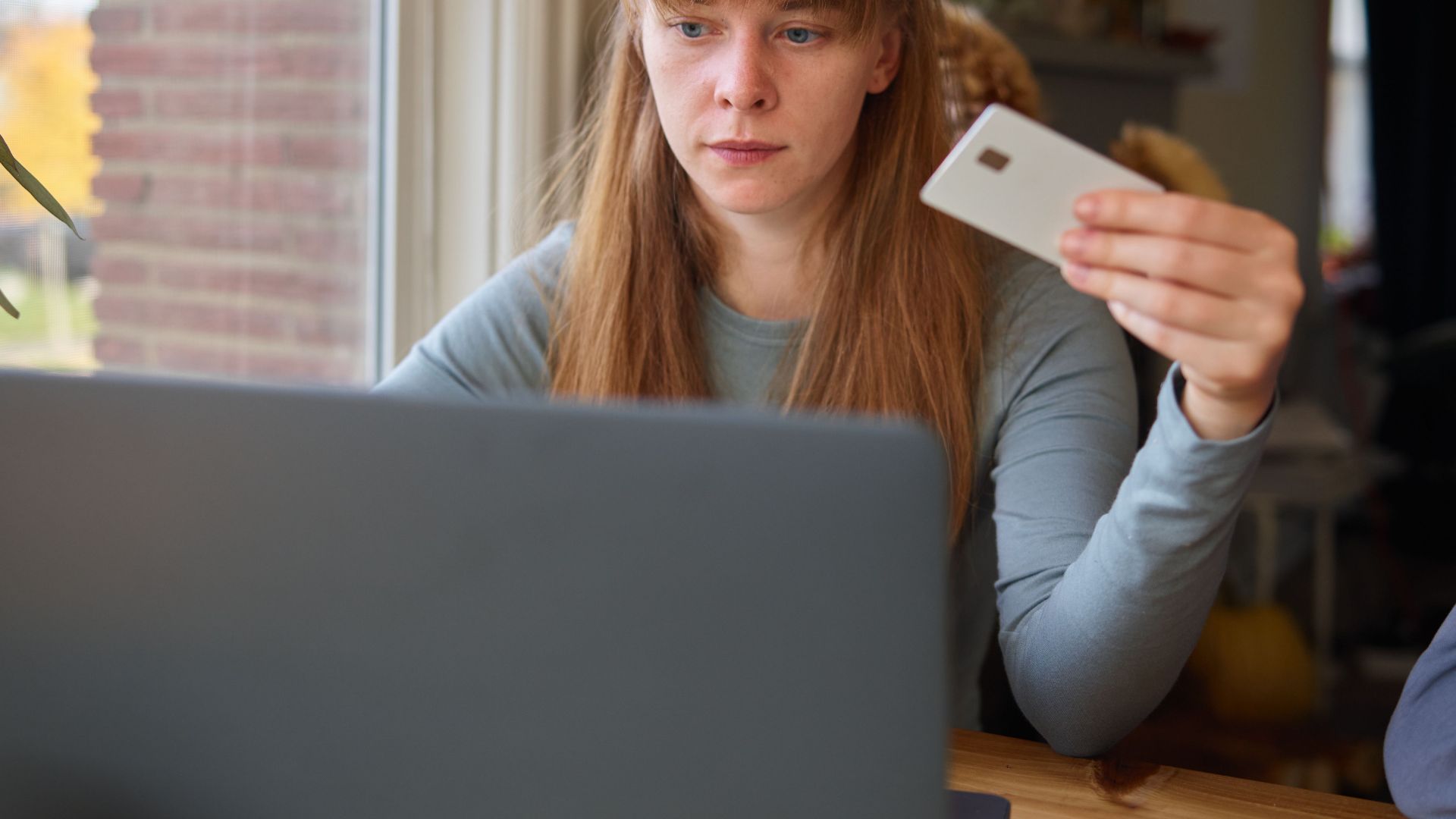 A young woman holds a credit card while using her laptop indoors.