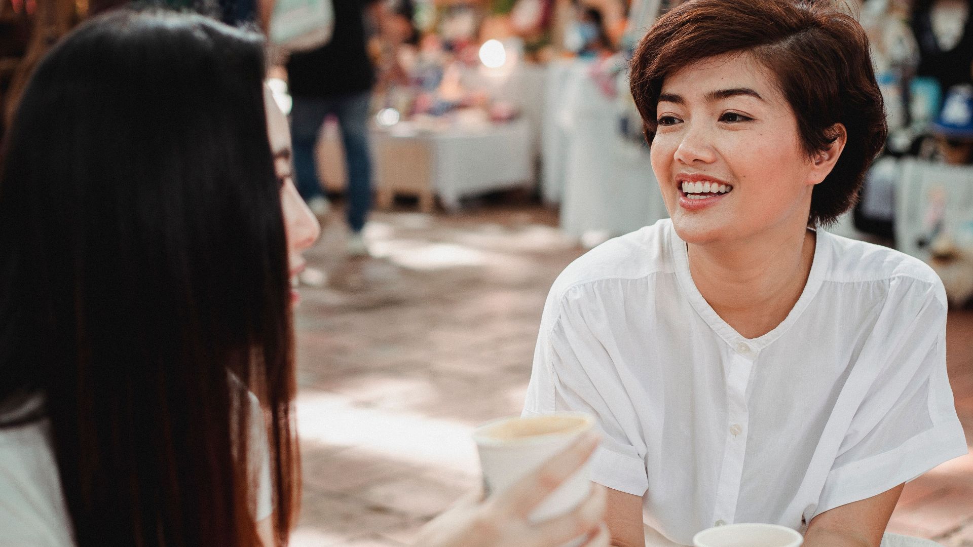 Happy young ethnic females having hot drinks and talking while sitting in outdoor cafeteria and looking at each other with smiles