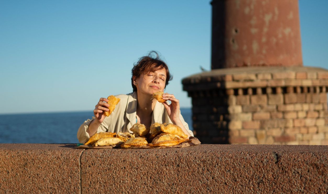 Adult woman savoring pastries outdoors near a stone structure by the sea, under bright day light