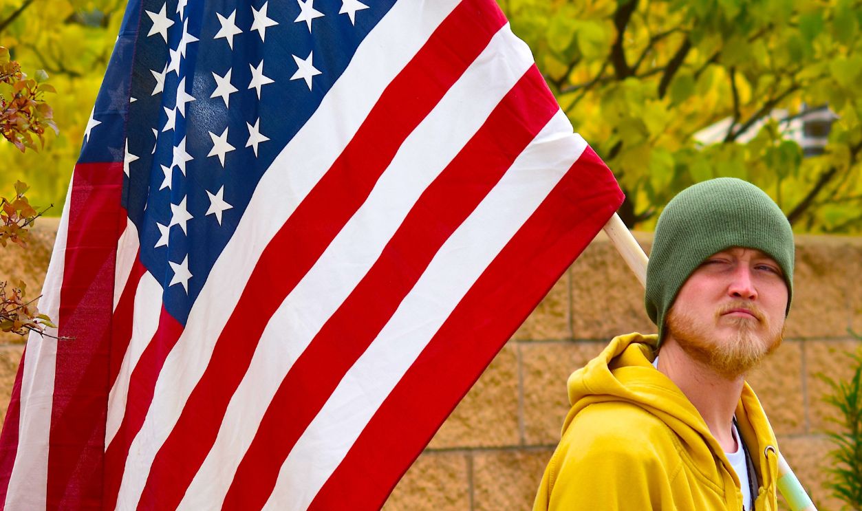 A young adult holding a large American flag, flashing a peace sign at an outdoor protest