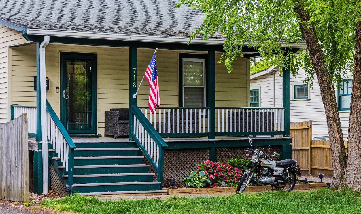 A quaint house with a porch, American flag, and motorcycle parked in the green yard