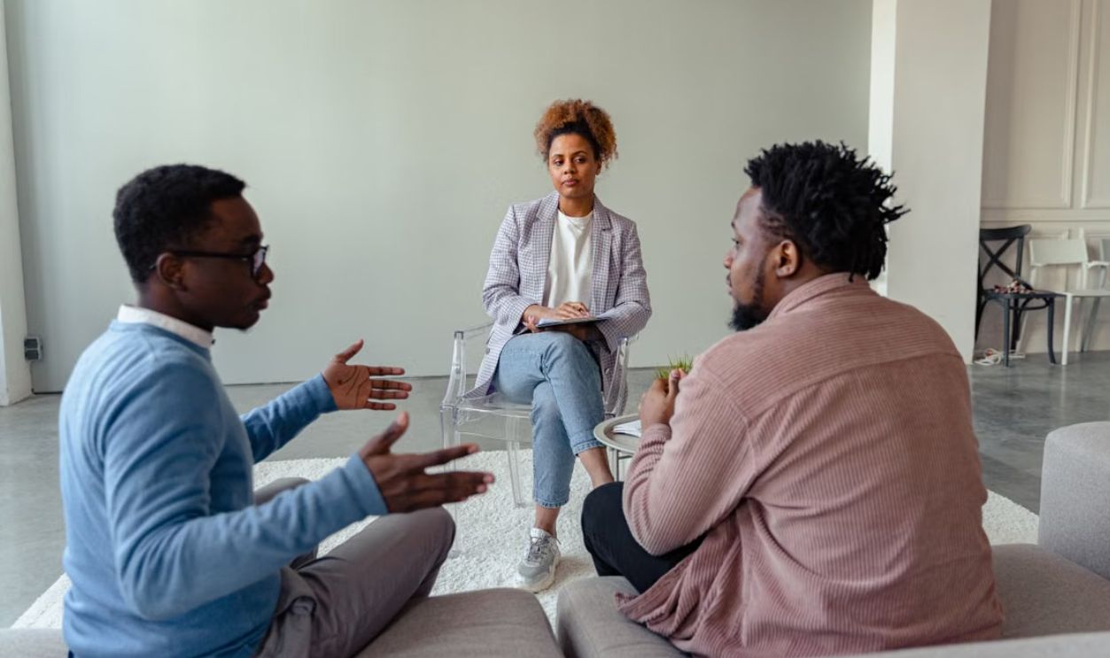 A therapist mediates a session with two men on a couch discussing mental health