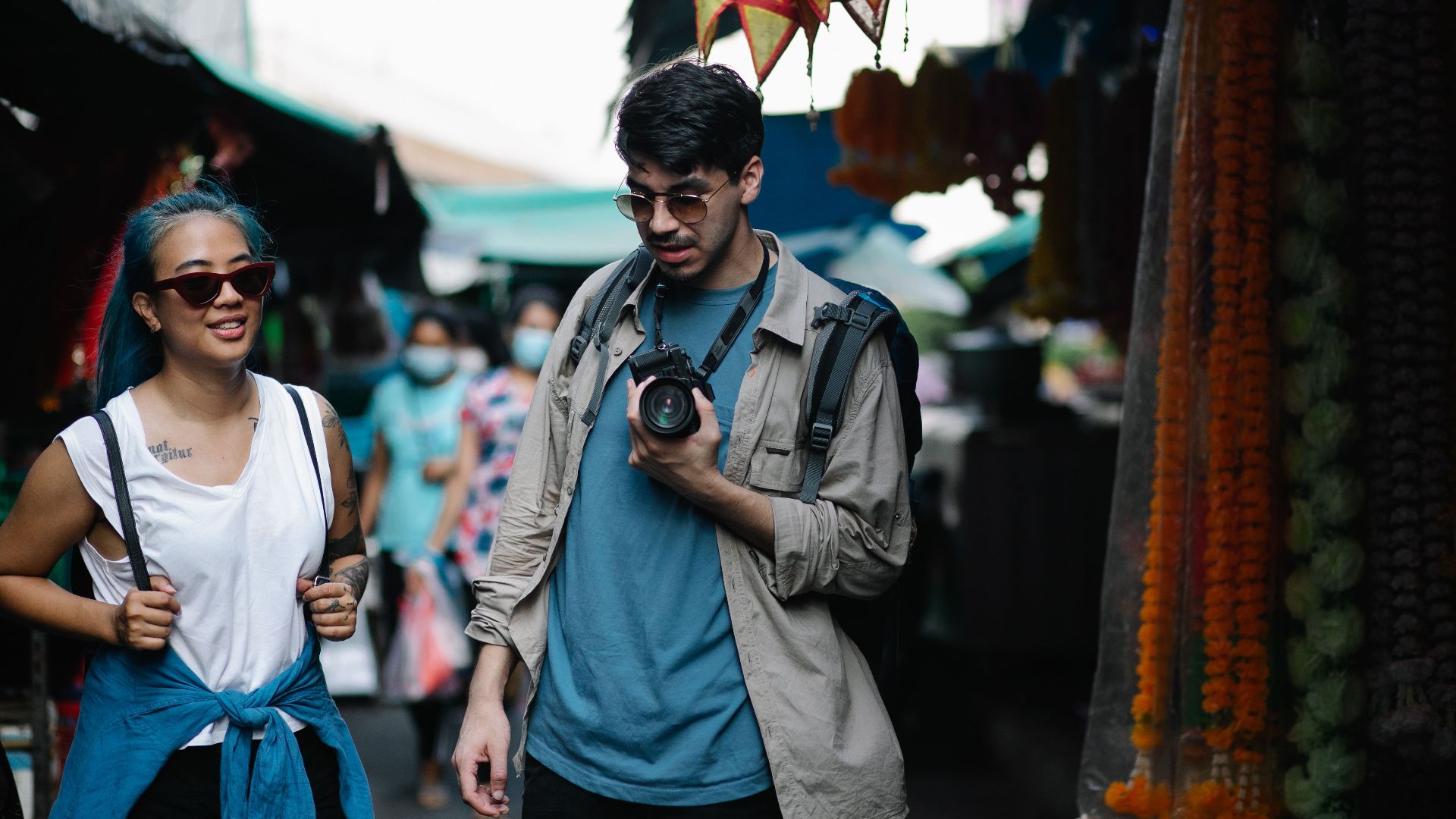 Two friends explore a bustling street market, capturing moments with a camera.