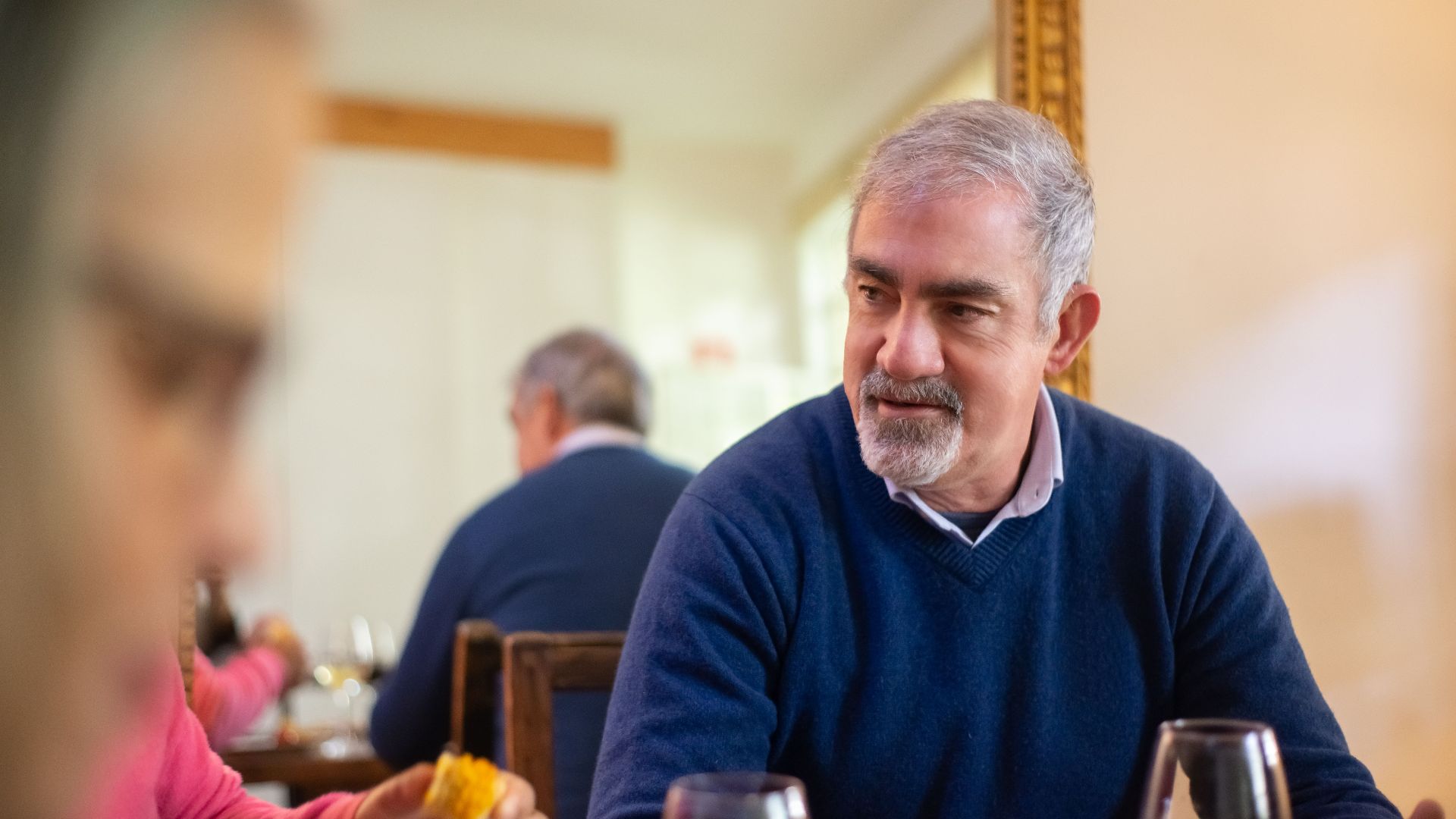 Senior man in a cozy indoor setting enjoying a meal with friends, wearing a blue sweater.