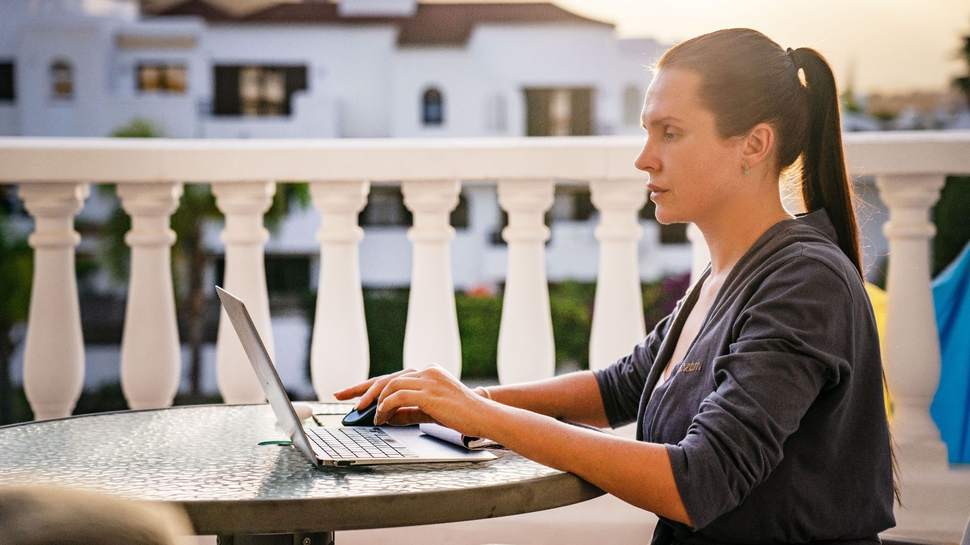 A focused woman works on her laptop outdoors on a sunny balcony, capturing a serene morning work vibe.