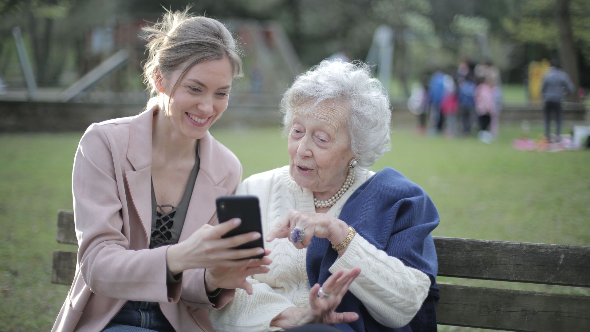 Delighted female relatives sitting together on wooden bench in park and browsing mobile phone while learning using