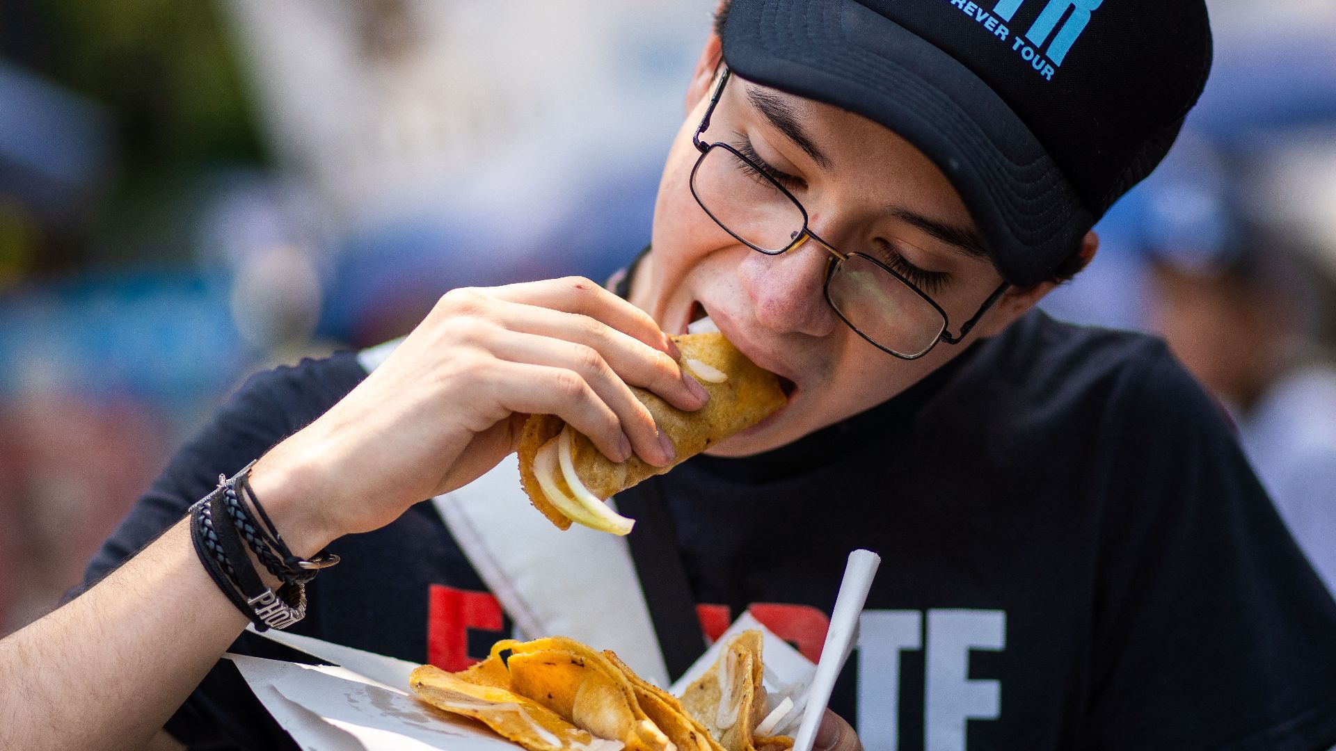 A young man wearing a cap and glasses enjoys street tacos in Mexico City.