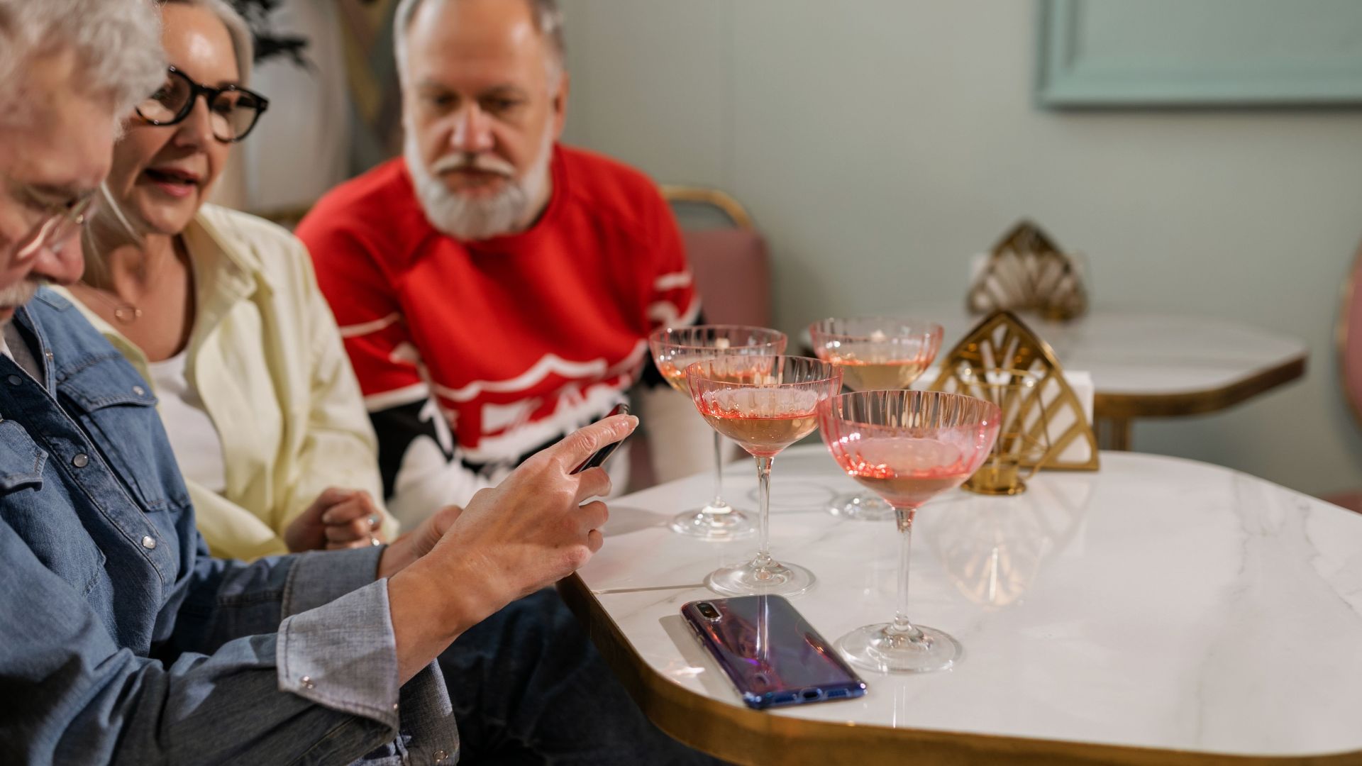 Three senior adults socializing at a cafe with cocktails, sharing a moment over a smartphone.