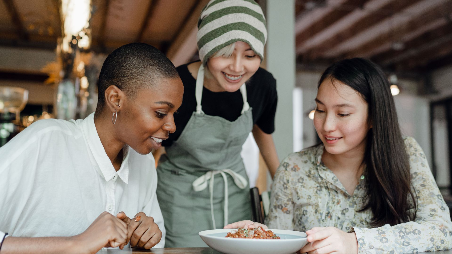 Smiling positive multiracial females in casual clothes sitting at table in modern cafe near cheerful waitress and looking at delicious dish