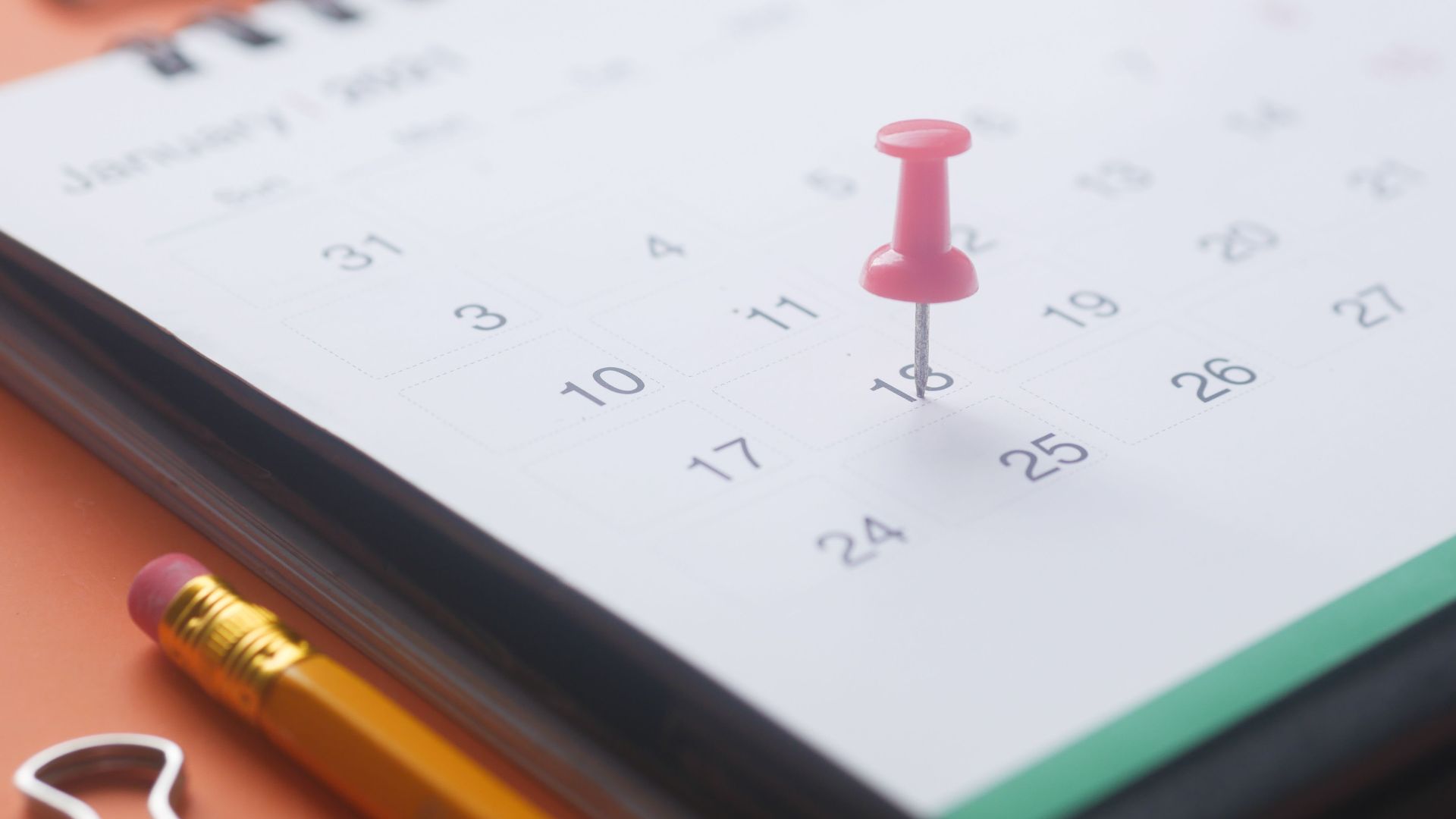 A close-up of a calendar page with a pink push pin, pencil, and binder clip on an orange background.