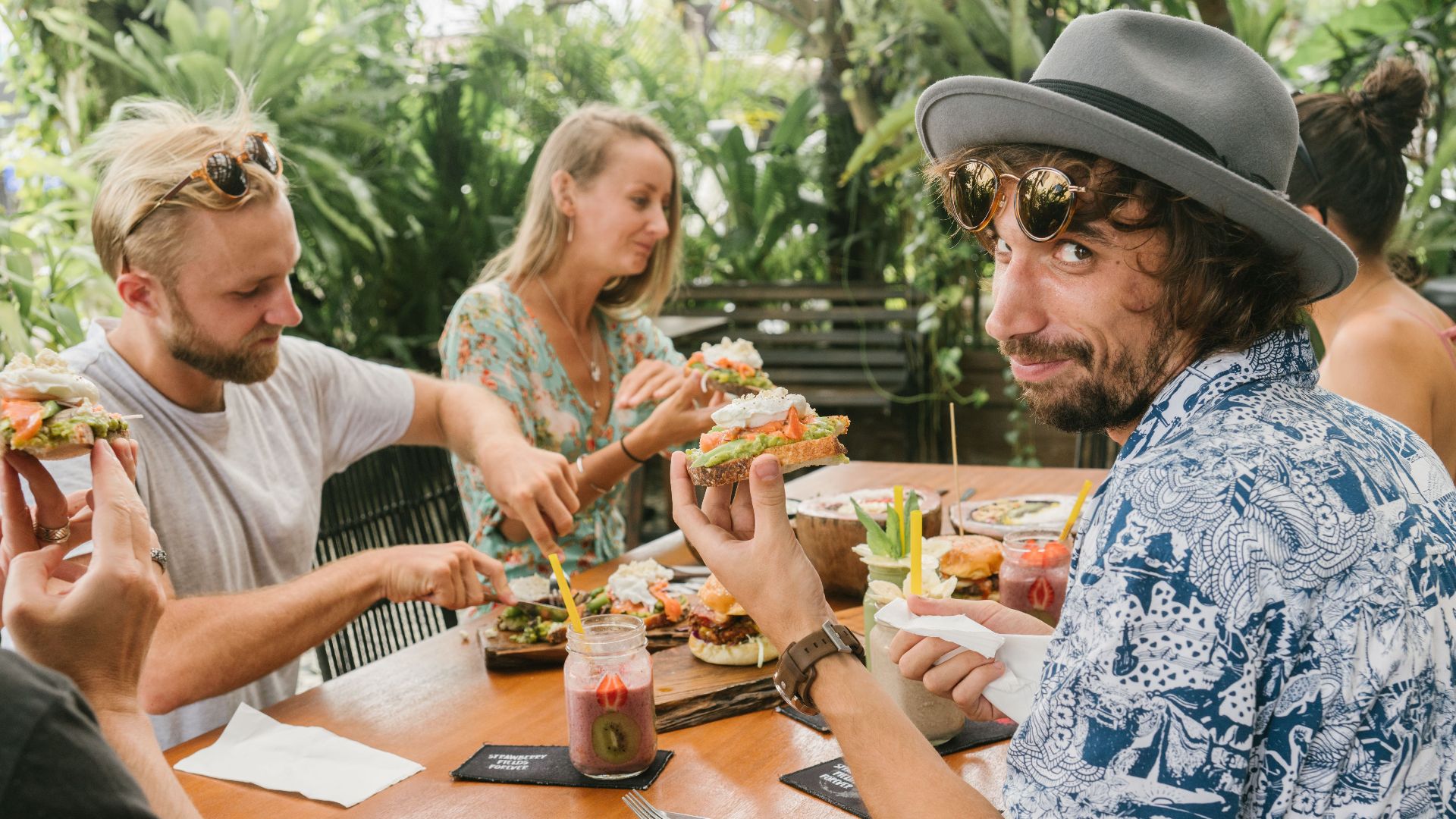 Friends gather around a table enjoying brunch outdoors with sandwiches and drinks.