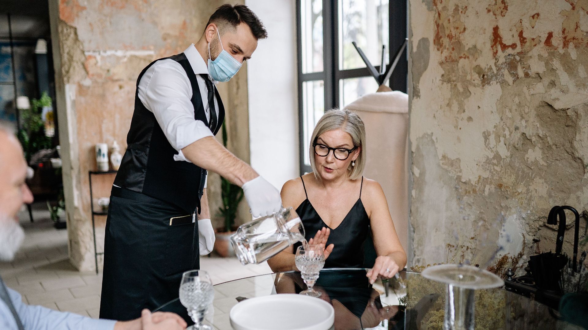 A waiter wearing a mask serves drinks to a seated couple in a stylish restaurant.