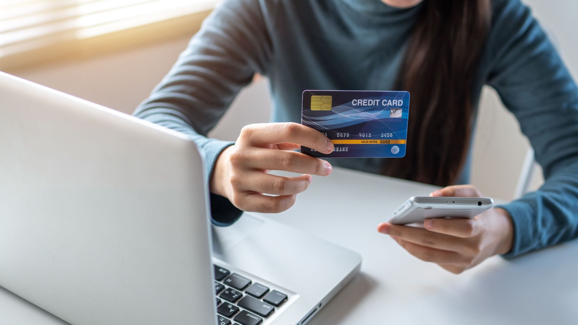 A woman uses a credit card and smartphone for online shopping on her laptop