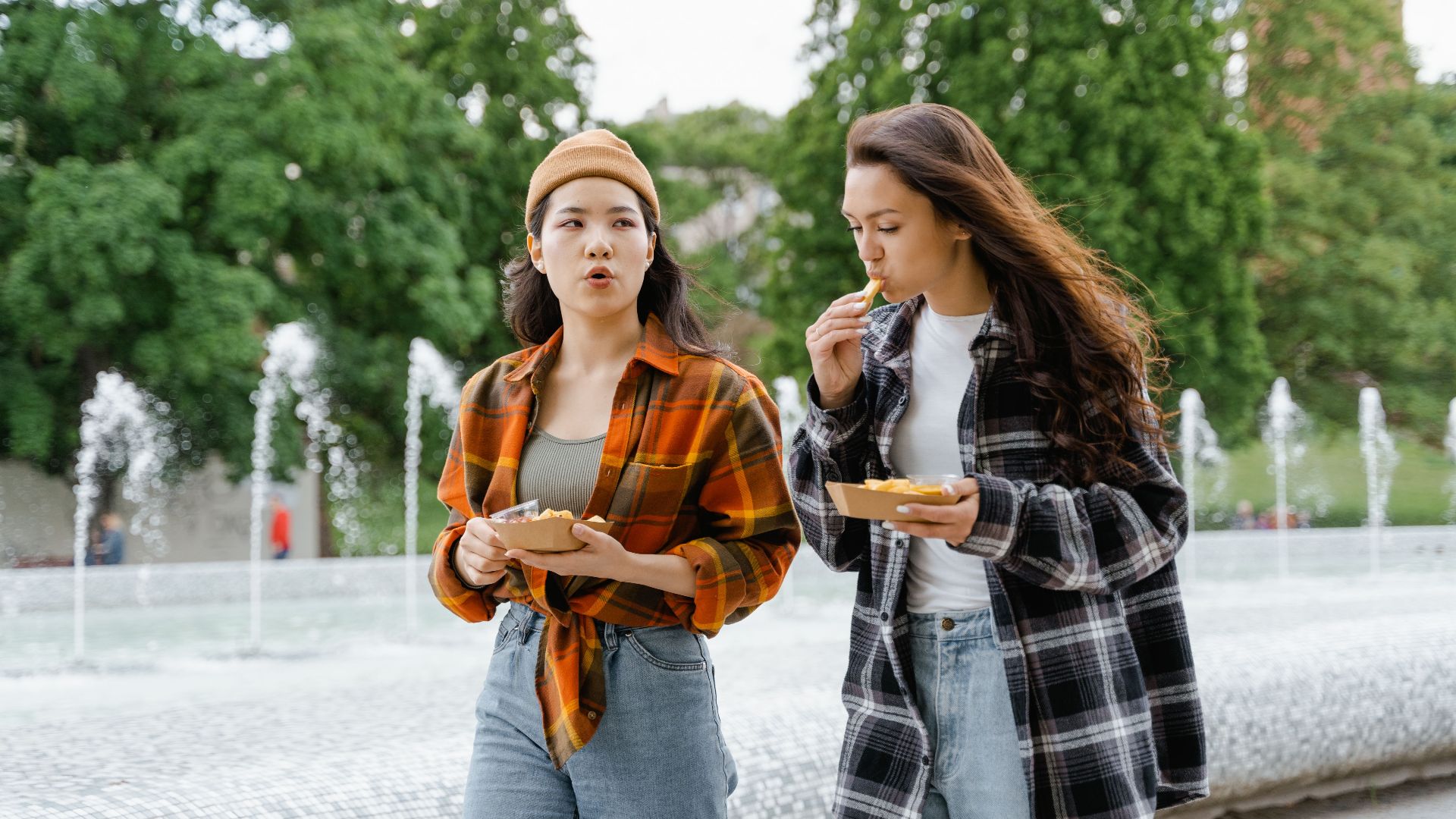 Young women eating snacks while walking in a park near a fountain on a sunny day.