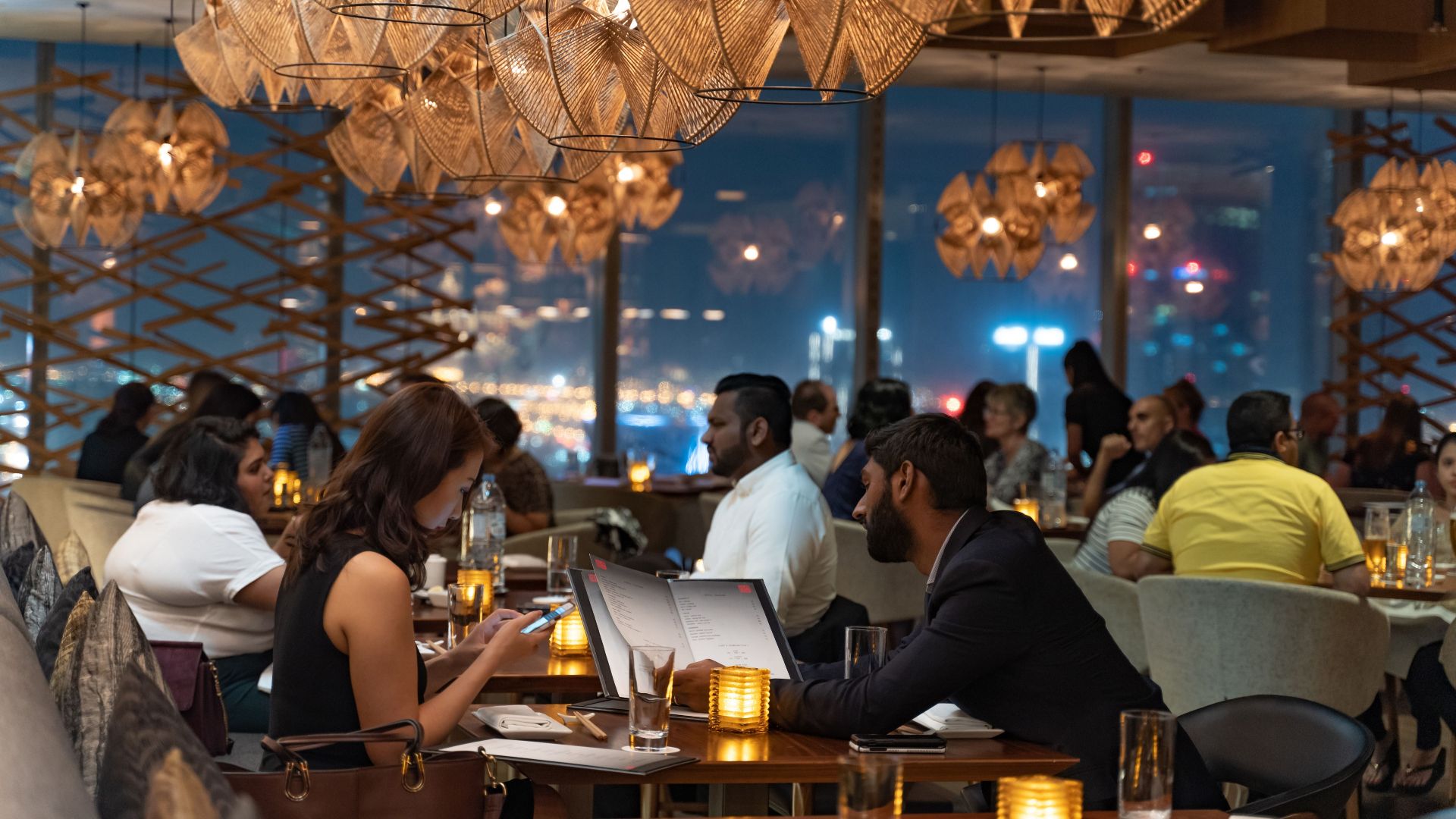 A stylish restaurant scene with people dining, featuring ambient lighting and a city view.