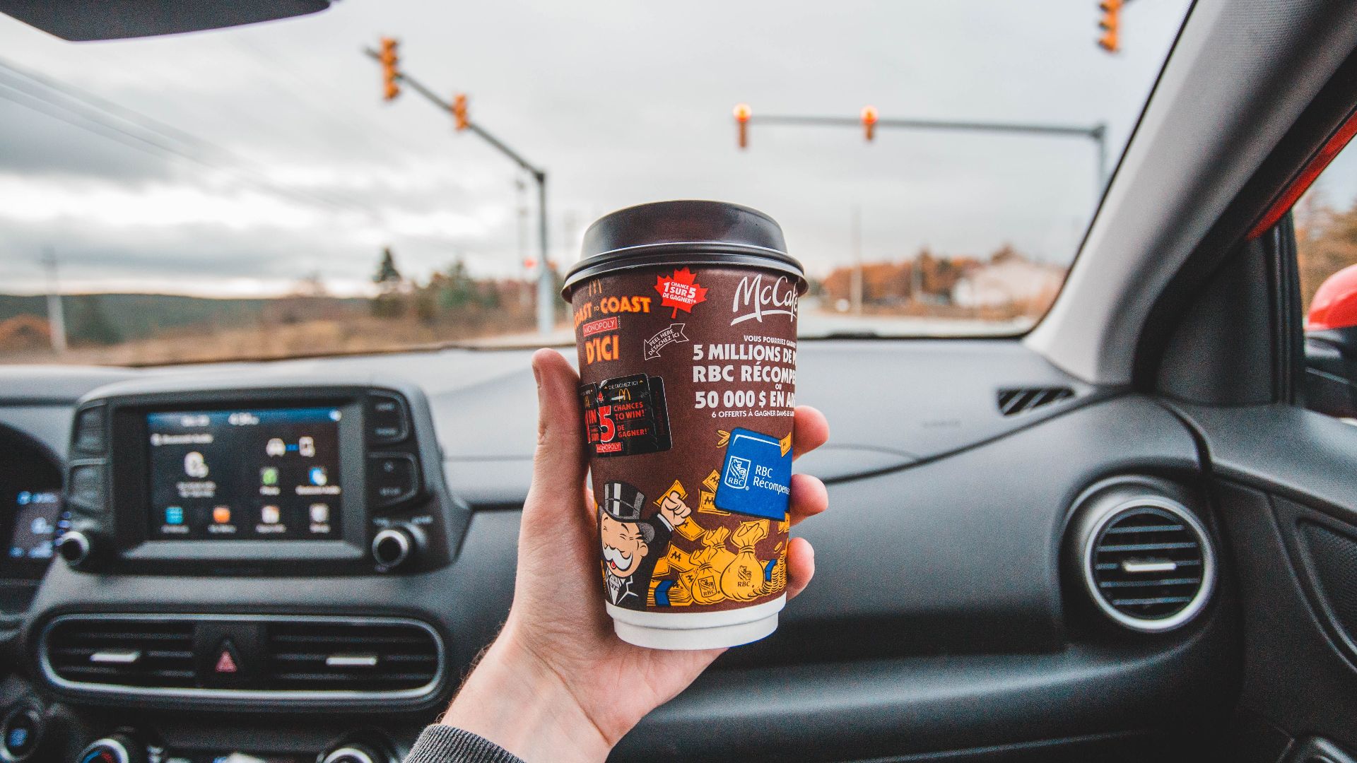 Hand holding coffee cup inside car with intersection view ahead. Captured in overcast weather.