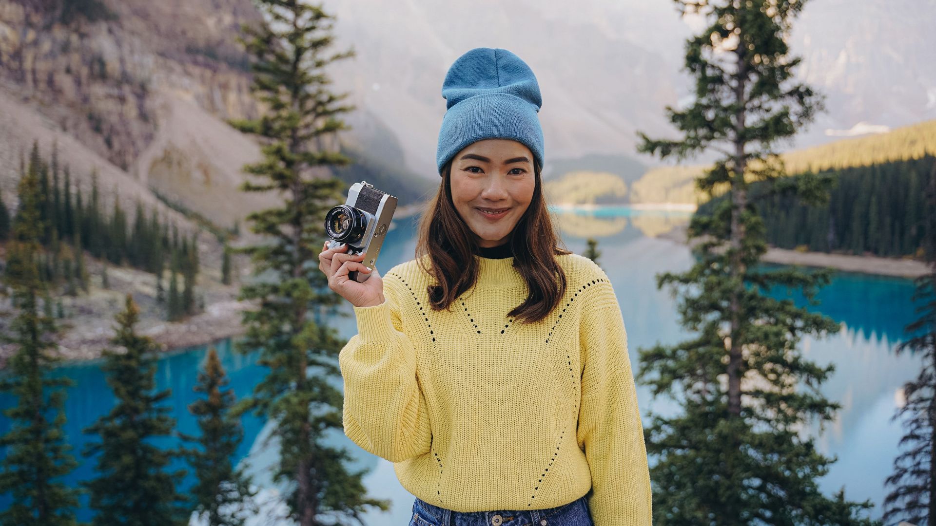 A young woman holds a camera, smiling by the vibrant blue waters of Peyto Lake, surrounded by mountains.