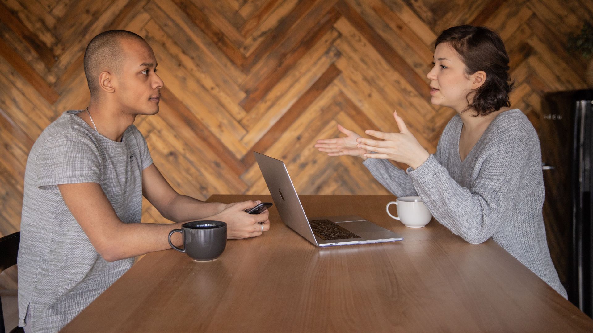 Focused woman explaining opinion to ethnic male coworker during business teamwork sitting at table with laptop and coffee cups in cozy kitchen against wooden wall and looking at each other