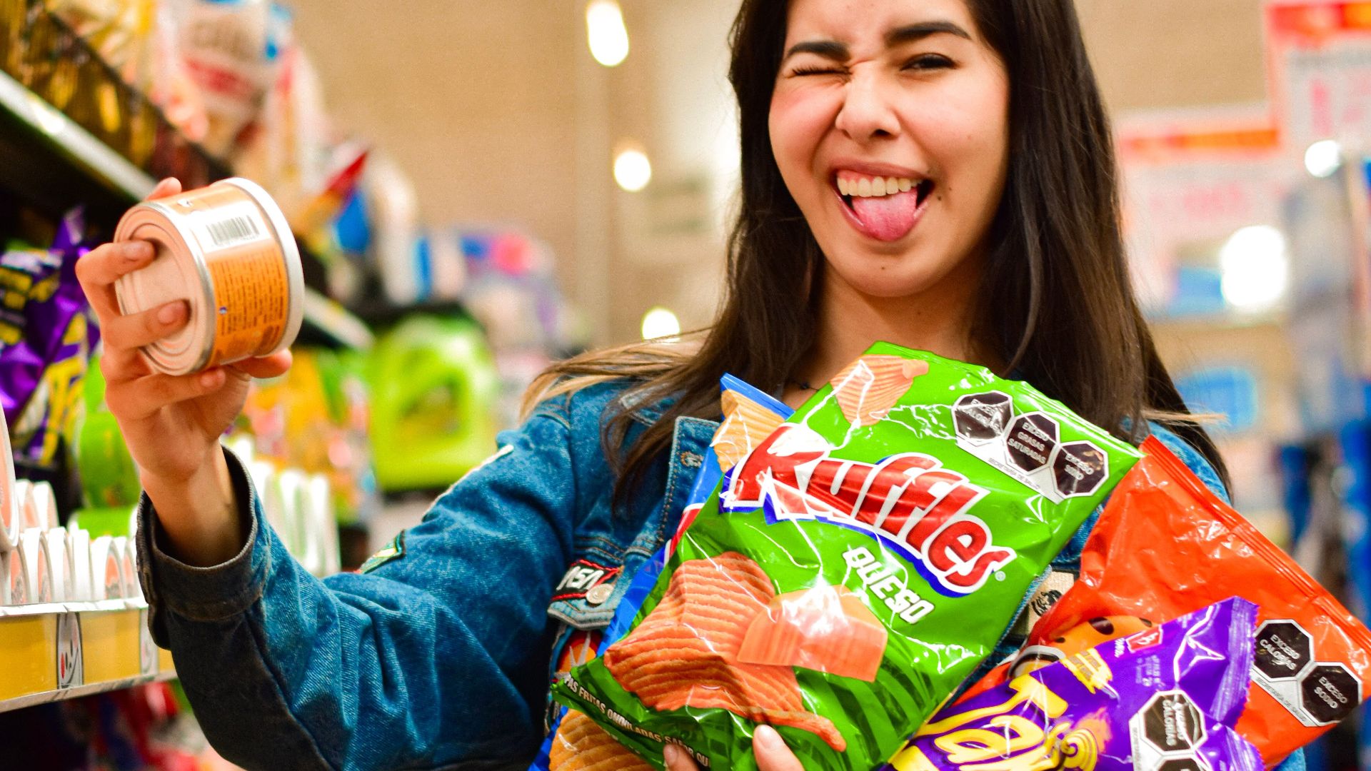 A happy woman holding snacks in a vibrant supermarket aisle, smiling playfully.
