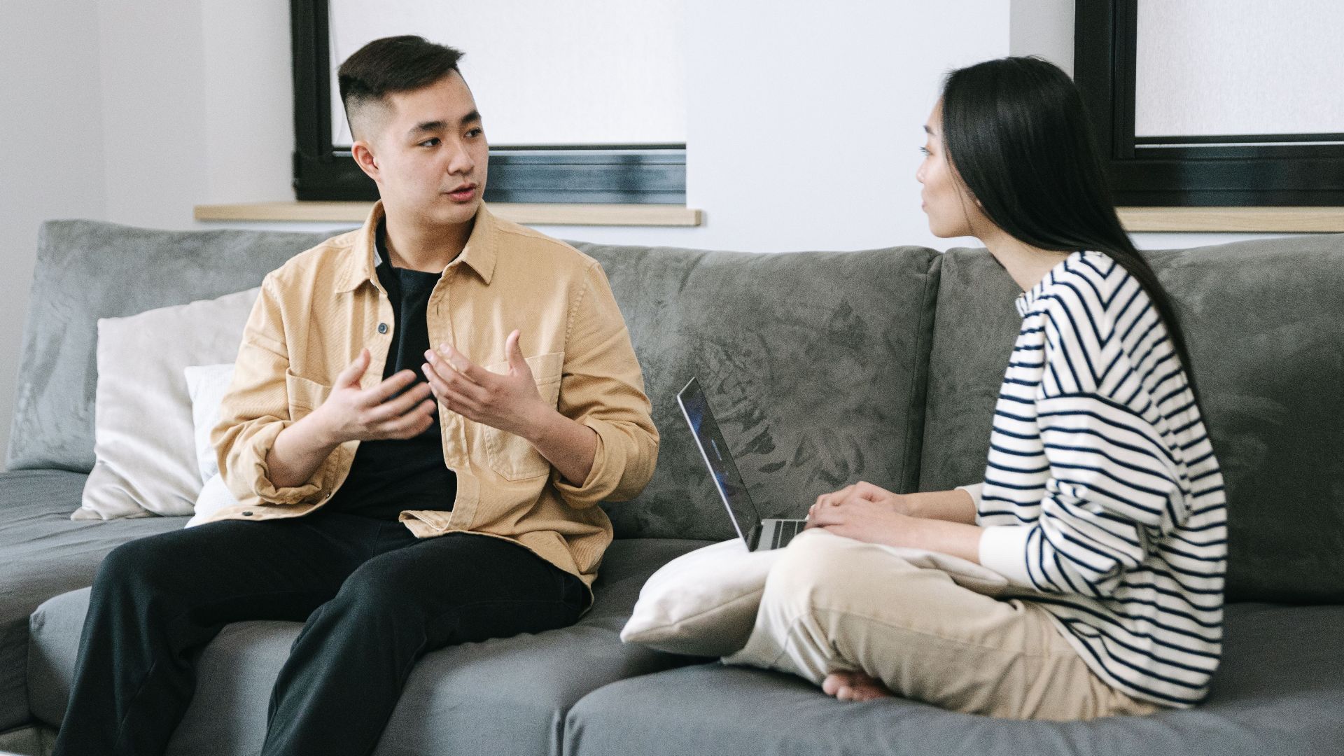 Two young adults engaged in a conversation, sitting on a couch at home.