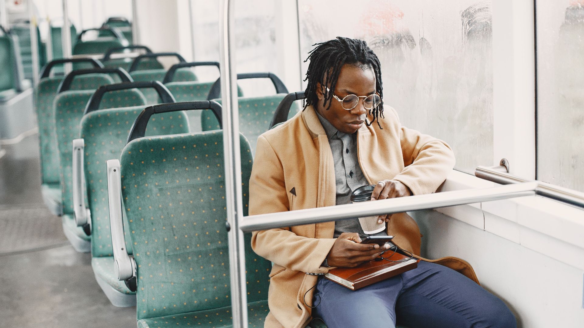 African American man on bus, wearing facemask and coat, using smartphone for commuting.