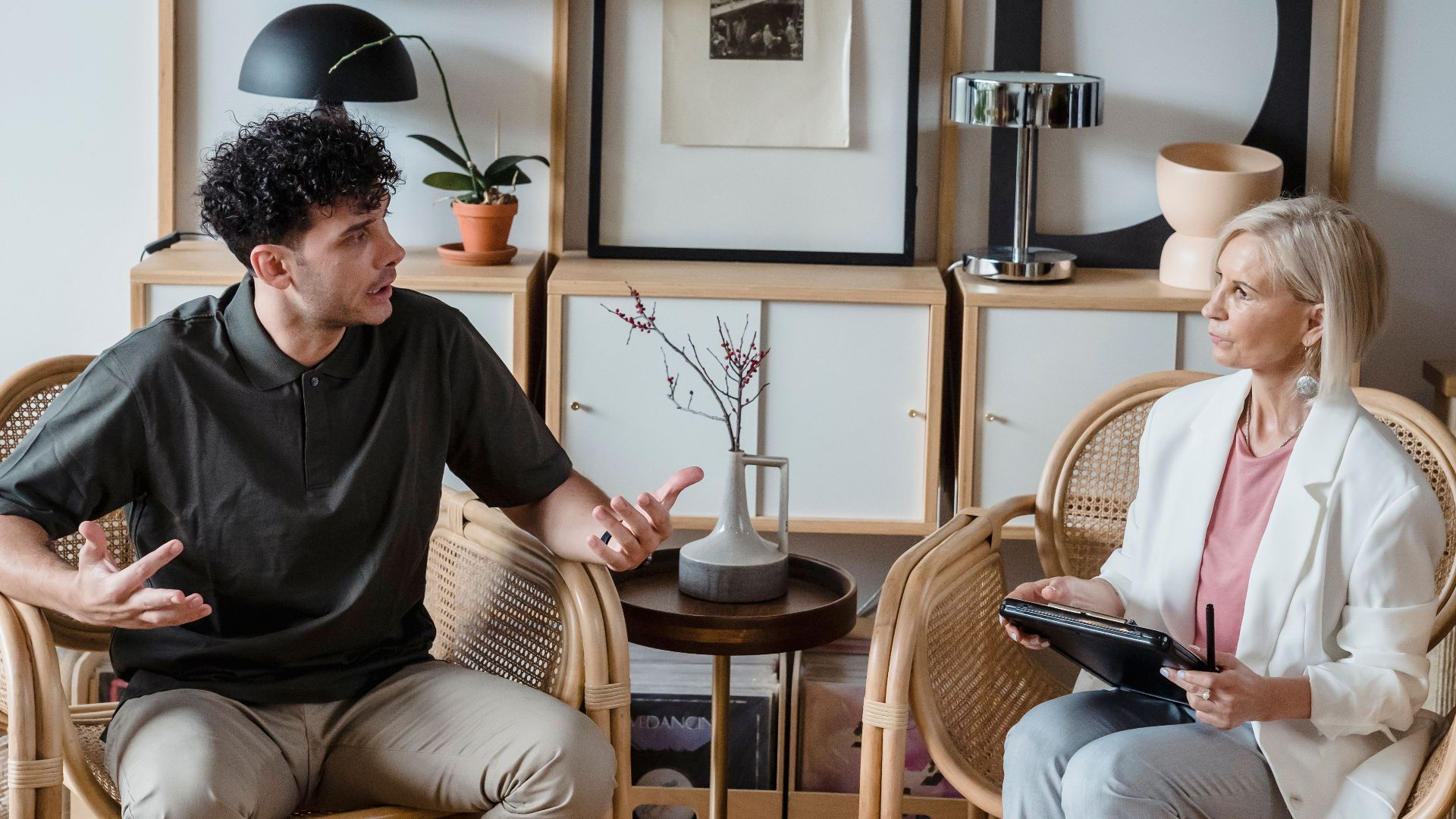 A man and woman in discussion in a modern living room with wooden chairs and artistic decor.