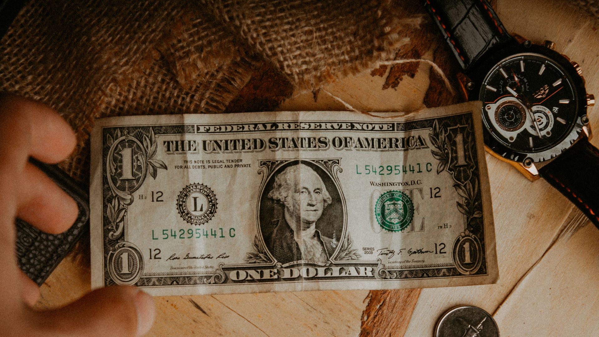 A person reaches for a one dollar bill surrounded by coins and a watch on a wooden table.
