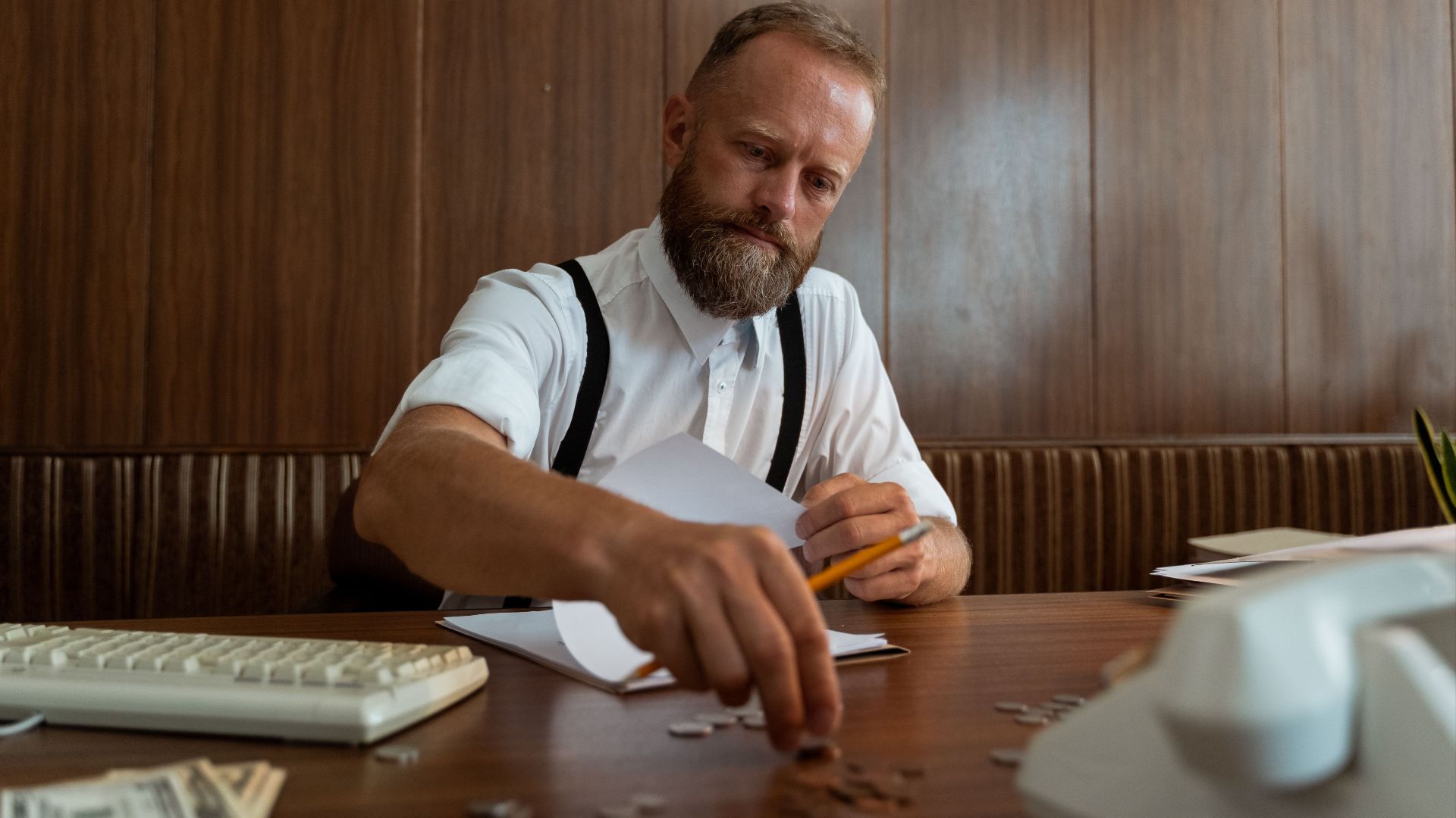 A bearded businessman wearing suspenders counts coins at a vintage workspace, embodying retro office style.